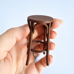 May include: A miniature brown stool featuring a circular seat, four curved legs, and a circular support ring connecting the legs. The stool is constructed with a smooth, matte finish and exhibits a classic bentwood-style design structure.