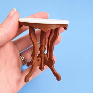 May include: A miniature round table with a smooth white top and a brown base featuring four curved legs and a central pedestal. The furniture piece is designed with intricate detailing on the support structure and stands on a flat base.