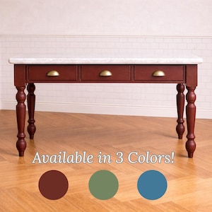 May include: A burgundy-colored desk with a white marble top and three drawers with brass handles. The desk has turned legs and is displayed on a hardwood floor. The text "Available in 3 Colors!" is below the desk.