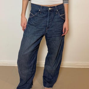 Könnte beinhalten: Dunkelblaue Jeans mit lockerer Passform. Die Jeans hat einen Knopfverschluss, Vordertaschen und einen leicht ausgefransten Saum. Die Person trägt silberne Loafer und eine gestreifte schwarz-weiße Jacke.