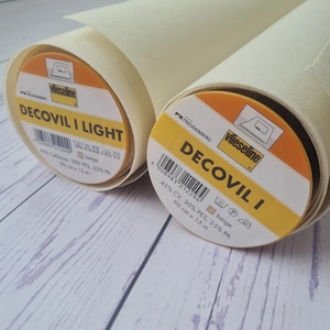 May include: Two rolls of beige Decovil I Light fabric, each with a label displaying the brand name, product details, and a barcode. The fabric is 90 cm wide and 15 m long.