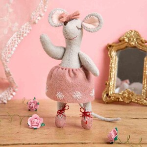 May include: A gray felt mouse doll wearing a pink tutu with white flower accents and pink ballet slippers tied with red ribbons. The mouse has a pink bow and is posed with one arm raised. Decorative roses and a gold mirror are in the background.
