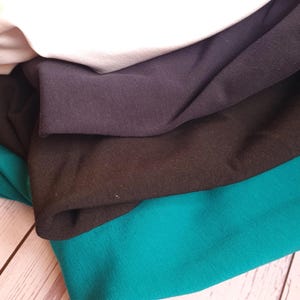 May include: A close-up of several fabric swatches in various colors. The colors include white, light purple, dark purple, black, and teal. The fabric appears to be a soft knit material, with visible folds and textures.