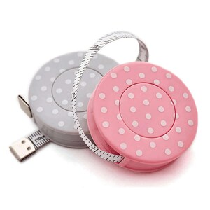 Op de afbeelding: Twee meetlinten met stippen. Eén is lichtgrijs met witte stippen en de andere is roze met witte stippen. De tape loopt uit de grijze rolmaat en toont metingen in centimeters tot 150 cm.