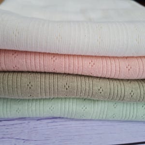May include: Four folded fabrics in pastel shades: white, pink, olive green, and light green. Each fabric has a ribbed texture with small, evenly spaced diamond-shaped perforations. The fabrics are stacked on a light purple surface.