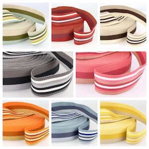 Può includere: Una collezione di rotoli di fettuccia a righe colorate. I rotoli mostrano varie combinazioni di colori, tra cui verde, rosso, grigio, rosa, arancione, blu e giallo, ciascuno con strisce a contrasto. La fettuccia è adatta per cinghie, cinture o scopi decorativi.