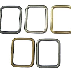 Könnte beinhalten: Fünf quadratische Metallringe in verschiedenen Ausführungen: Dunkelgrau, Silber, Gold, Antikmessing und Gunmetal. Diese Ringe werden wahrscheinlich für die Herstellung von Taschen, Riemen oder andere Bastelprojekte verwendet. Die Ringe werden vor weißem Hintergrund gezeigt.