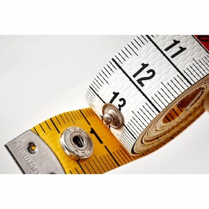 Puede incluir: Una cinta métrica amarilla y blanca con marcas y números negros. La cinta está parcialmente enrollada, mostrando los números 11, 12 y 13 pulgadas (28, 30 y 33 cm). La cinta tiene un extremo metálico con la palabra "Germany" impresa.