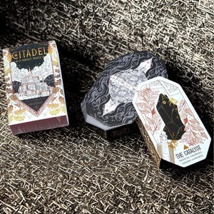 May include: Three oracle card decks with unique designs. The Citadel deck features a fantasy cityscape illustration. The Catalyst deck has a black crystal graphic. The third deck is black with a white city map design. All decks have gold accents.
