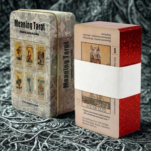 Könnte beinhalten: Ein Meaning Tarot Kartenspiel in einer dekorativen Dose. Die Dose ist beige mit den Worten "Meaning Tarot" und "Keywords, Reversed, Zodiac" aufgedruckt. Das Deck ist mit einem weißen Band umwickelt und hat rote Kanten.