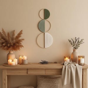 Puede incluir: Una consola de madera con velas, jarrones y decoración de pared. El arte mural presenta tres diseños circulares con hilo verde y blanco. La mesa está decorada con velas encendidas, flores secas y una manta beige.
