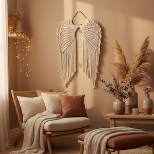 Op de afbeelding: Een beige macramé engelenvleugel wanddecoratie wordt getoond boven een rotan stoel met kussens en een deken. Een kleine houten tafel bevat vazen met gedroogde planten. De kamer heeft een neutraal kleurenpalet.