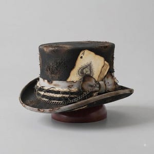 May include: A distressed black top hat featuring a weathered ace of spades playing card tucked into a white band. The hat is decorated with a small bow, a thin metal chain, and frayed rope detailing, creating a worn and textured appearance.