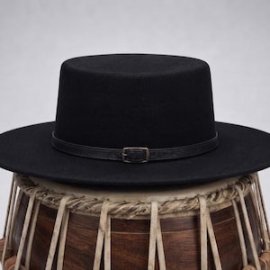 Puede incluir: Un sombrero negro de ala plana con copa plana y una fina correa de cuero negro con una pequeña hebilla metálica alrededor de la base. El sombrero está colocado sobre un tambor de madera con cordones de color claro visibles en el cuerpo.