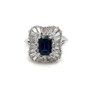 Puede incluir: Un anillo plateado con una piedra preciosa rectangular de color azul oscuro en el centro, rodeada por un halo de piedras preciosas más pequeñas y claras. El diseño del anillo incluye un patrón radiante en forma de sol alrededor de la piedra central.