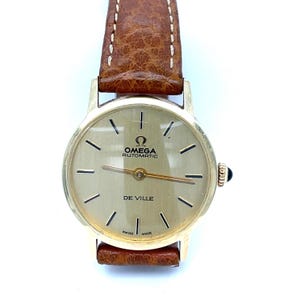 May include: A gold-toned Omega De Ville automatic wristwatch with a brown leather strap. The watch face is a light gold color with black markers and hands. The watch has the Omega logo and the words "AUTOMATIC" and "DE VILLE" on the face. "SWISS MADE" is printed at the bottom.