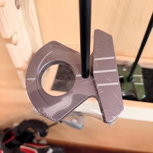 Könnte beinhalten: Ein neuer Golf-Putter mit einem dunkelgrauen Kopf und einem schwarzen Schaft. Der Putterkopf hat drei weiße Ausrichtungslinien und ist noch in Plastik verpackt. Entwickelt für Präzision und Kontrolle auf dem Grün.
