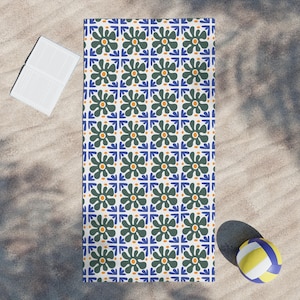 May include: A rectangular beach towel with a repeating floral pattern in green, blue, and orange on a white background. A white book and a yellow, blue, and white volleyball are on the sand next to the towel.
