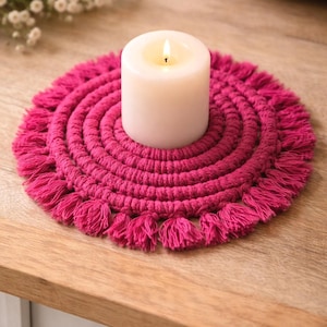 May include: A lit white pillar candle sits on a round, hot pink macrame coaster with fringe. The coaster is made of thick, woven material. The candle and coaster are on a light wood surface.