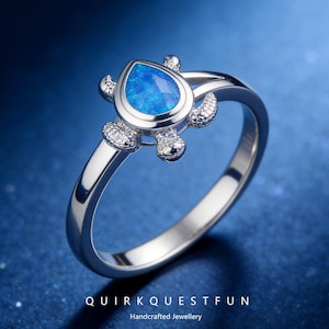 May include: A silver ring with a turtle design, featuring a blue, teardrop-shaped stone. The turtle's head and flippers are decorated with small, clear stones. The ring is set against a blue background, with the text "QUIRKQUESTFUN" and "Handcrafted Jewellery".
