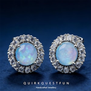 May include: A pair of stud earrings featuring a central, round, light blue opal stone surrounded by a halo of small, clear, round gemstones. The earrings are set in a silver-colored metal. The text "QUIRK QUEST FUN" and "Handcrafted Jewellery" are at the bottom.