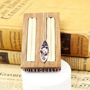 May include: A silver necklace featuring a surfboard pendant with a turtle design. The turtle has a pink and white opal-like center. The pendant is on a silver chain and displayed on a wooden jewelry holder.