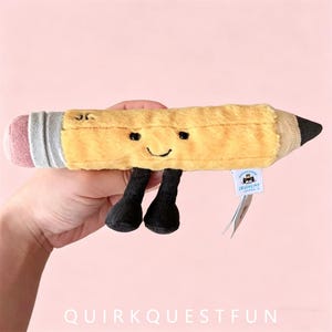 May include: A plush pencil toy featuring a yellow body, a pink eraser end, a grey metal-colored band, and a dark brown pointed tip. The pencil has a stitched smiling face with black eyes and two small black corduroy legs dangling below.