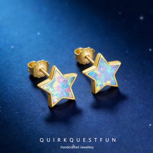 May include: A pair of gold-colored star-shaped stud earrings with iridescent opal-like centers. The earrings are set against a dark blue background. The text "QUIRKQUESTFUN Handcrafted Jewellery" is at the bottom.