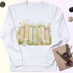 Puede incluir: Sudadera blanca con una ilustración caprichosa de conejitos cuidando flores y libros. El diseño incluye libros en colores pastel, margaritas y adorables conejitos, creando una estética encantadora con temática primaveral. La sudadera es una prenda cómoda e informal.