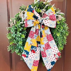 Op de afbeelding: Een ronde krans van groen buxus, versierd met een grote, veelkleurige stoffen strik. De strik heeft een patchworkontwerp in rood, wit, blauw en geel, met motieven van sterren, bloemen, klokken en kleine Amerikaanse vlaggetjes op de stof.