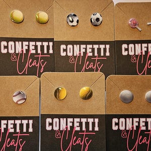 May include: A collection of stud earrings featuring sports ball designs, including softballs, baseballs, soccer balls, pickleball paddles, and volleyballs. The earrings are displayed on brown cards with the text "Confetti & Cleats" in white and pink.