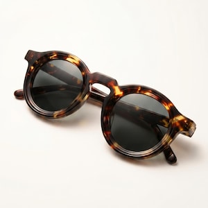 May include: Round sunglasses in a tortoiseshell design. The frames are brown and yellow, and the lenses are dark. These sunglasses have a classic, vintage style.