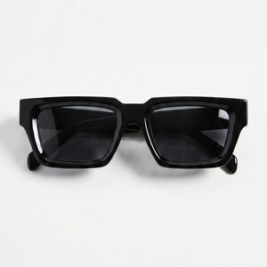 May include: Black sunglasses with a rectangular frame. The sunglasses have dark lenses and a thick, glossy black frame. The temples are also black and have a slight curve. The sunglasses are set against a white background.