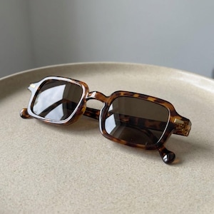 Puede incluir: Gafas de sol cuadradas con estampado de carey. Las gafas de sol tienen lentes oscuras y una montura marrón y beige. Las gafas están sobre una superficie de color claro.