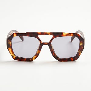 May include: Geometric sunglasses in a tortoiseshell design. The frames have an angular shape with a distinctive bridge cutout. The lenses are a solid grey colour, providing a modern and stylish appearance.
