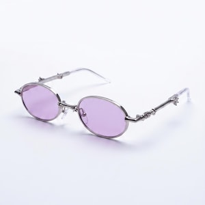 Peut inclure: Lunettes de soleil ovales avec une monture argentée et des verres violets. Les branches présentent des ornements décoratifs. Les lunettes sont posées sur un fond blanc.