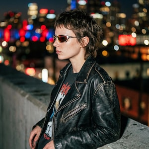K&ouml;nnte beinhalten: Eine Person tr&auml;gt eine schwarze Lederjacke und Sonnenbrille vor einer Stadtkulisse. Die Jacke hat einen Rei&szlig;verschluss und einen Used-Look. Die Sonnenbrille hat einen silbernen Rahmen und rot get&ouml;nte Gl&auml;ser.