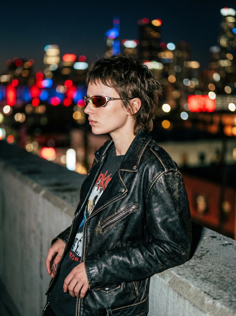 K&ouml;nnte beinhalten: Eine Person tr&auml;gt eine schwarze Lederjacke und Sonnenbrille vor einer Stadtkulisse. Die Jacke hat einen Rei&szlig;verschluss und einen Used-Look. Die Sonnenbrille hat einen silbernen Rahmen und rot get&ouml;nte Gl&auml;ser.