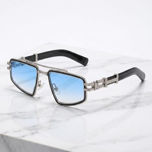 Peut inclure: Lunettes de soleil avec une monture en métal argenté et un double pont distinctif. Les verres présentent un dégradé bleu, passant d'un bleu foncé en haut à une teinte plus claire en bas. Les branches sont noires avec des détails argentés.
