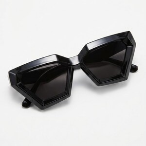 Peut inclure: Lunettes de soleil géométriques noires au design angulaire audacieux. Les lunettes présentent une monture noire épaisse et brillante et des verres foncés. La forme générale est un style moderne et avant-gardiste.