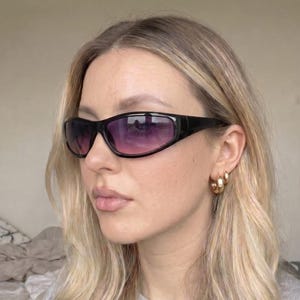 May include: Black sunglasses with a wraparound design and gradient purple lenses. The sunglasses have a thick, rectangular frame. The image shows a close-up of the sunglasses on a person.