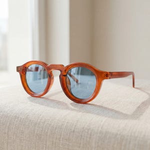 May include: Round, amber-colored sunglasses with blue lenses. The sunglasses are resting on a neutral-toned surface, with a window in the background. The frames have a classic, retro design.