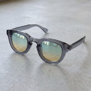 Peut inclure: Lunettes de soleil grises avec une monture carrée et des verres dégradés passant du jaune au bleu. Les lunettes de soleil ont un design moderne et minimaliste.