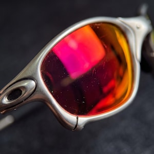 K&ouml;nnte beinhalten: Nahaufnahme einer Sonnenbrille mit silbernem Rahmen und leuchtend roten und orangen Gl&auml;sern. Die Gl&auml;ser reflektieren das Licht und erzeugen einen farbenfrohen Farbverlauf. Die Sonnenbrille hat ein schlankes, modernes Design.