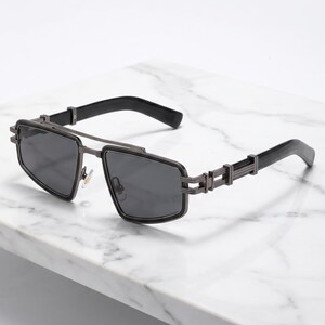 Peut inclure: Lunettes de soleil gris acier avec une monture géométrique angulaire et des verres teintés foncés. Les branches présentent un détail métallique segmenté de style industriel qui se connecte à des embouts en plastique noir lisse.