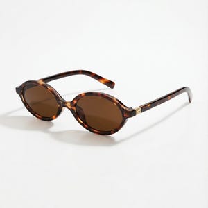 Könnte beinhalten: Ovale Sonnenbrille mit Schildpattmuster. Die Fassungen und Bügel haben ein dunkelbraunes und bernsteinfarbenes Design. Die Gläser sind einfarbig braun, und die Bügel haben einen goldfarbenen Akzent in der Nähe der Scharniere. Die Sonnenbrille ist vor weißem Hintergrund.