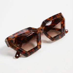 May include: Geometric-shaped sunglasses with a tortoiseshell pattern. The frames have a unique, angular design, and the lenses are tinted brown. The sunglasses are set against a white background.