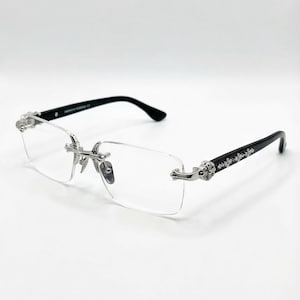 May include: Rimless spectacles with black arms and silver decorative accents. The lenses are rectangular and clear. The arms have a decorative floral design. The bridge and hinges are silver. The spectacles are on a white background.