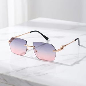 Peut inclure: Lunettes de soleil sans monture avec des verres rectangulaires présentant un dégradé passant du violet en haut au rose en bas. La monture est composée de métal doré fin avec des détails décoratifs aux charnières et des embouts noirs.