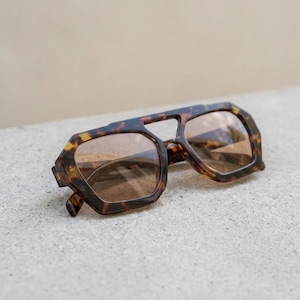Peut inclure: Lunettes de soleil à motif écaille de tortue avec un design géométrique. Les montures sont marron et beige, et les verres sont marron clair. Les lunettes sont posées sur une surface gris clair.